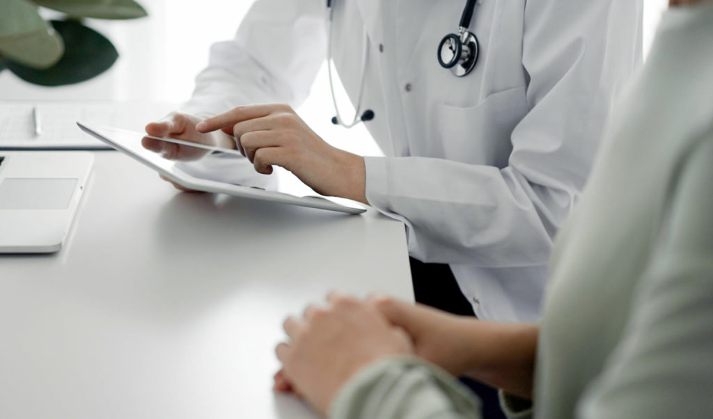 Médecin et patient assis au bureau de la clinique. L'accent est mis sur les mains d'une femme médecin utilisant une tablette, en gros plan.